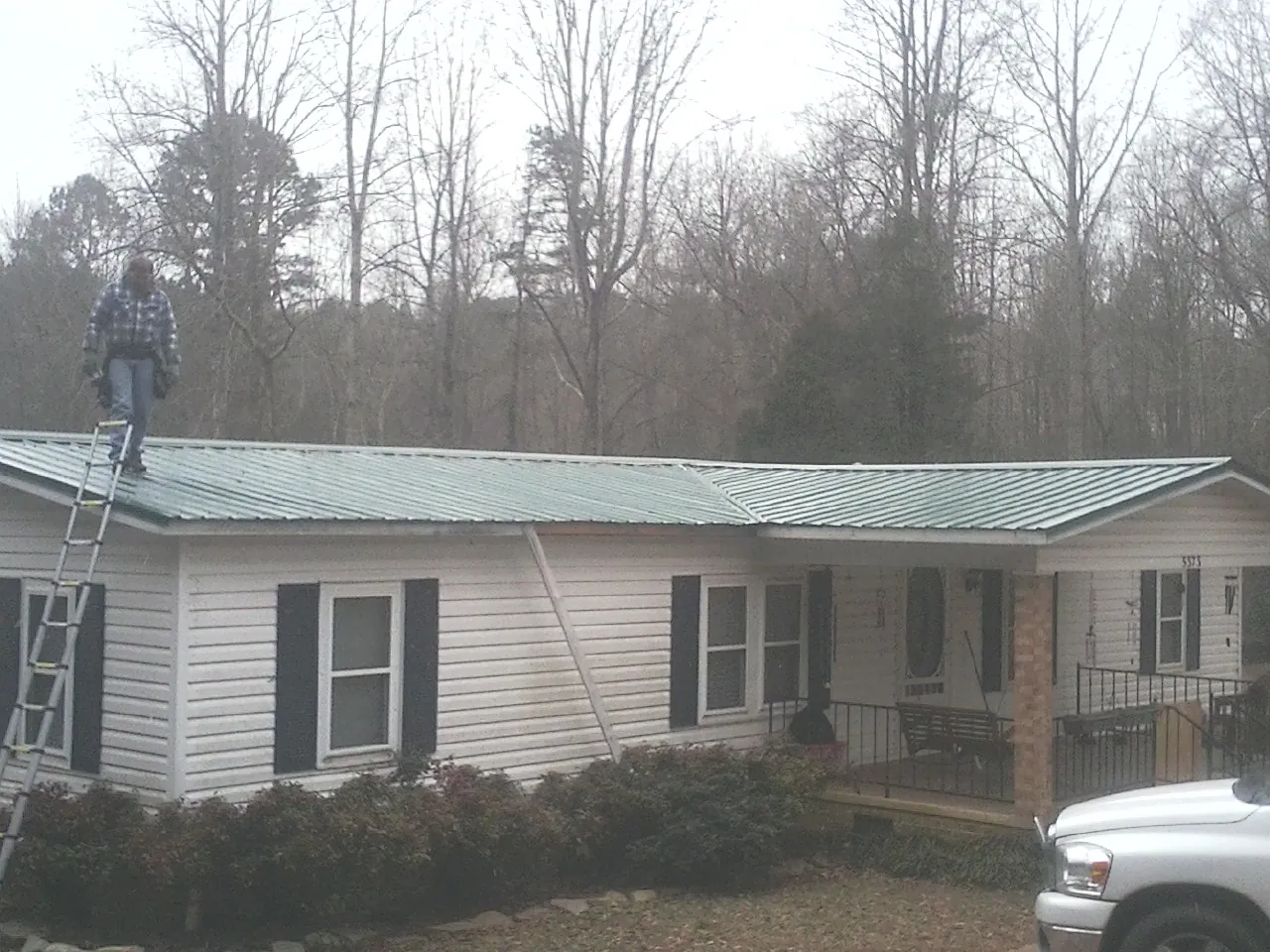 Worker on a ladder beside a new green metal roof on a ranch home in Cardinal