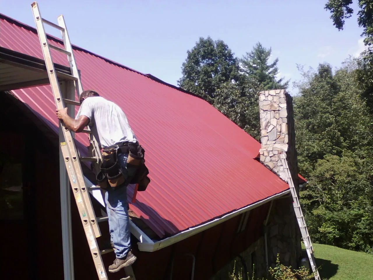 Roofer on a ladder beside a red metal roof and stone chimney — Soffit & Fascia Repair by Roofing Brothers in Cardinal